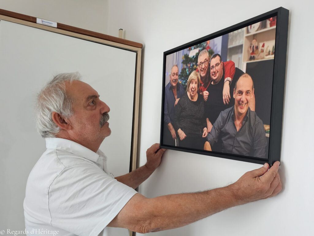 Un homme accroche au mur un tableau encadré représentant un portrait de famille de cinq personnes devant un sapin de Noël